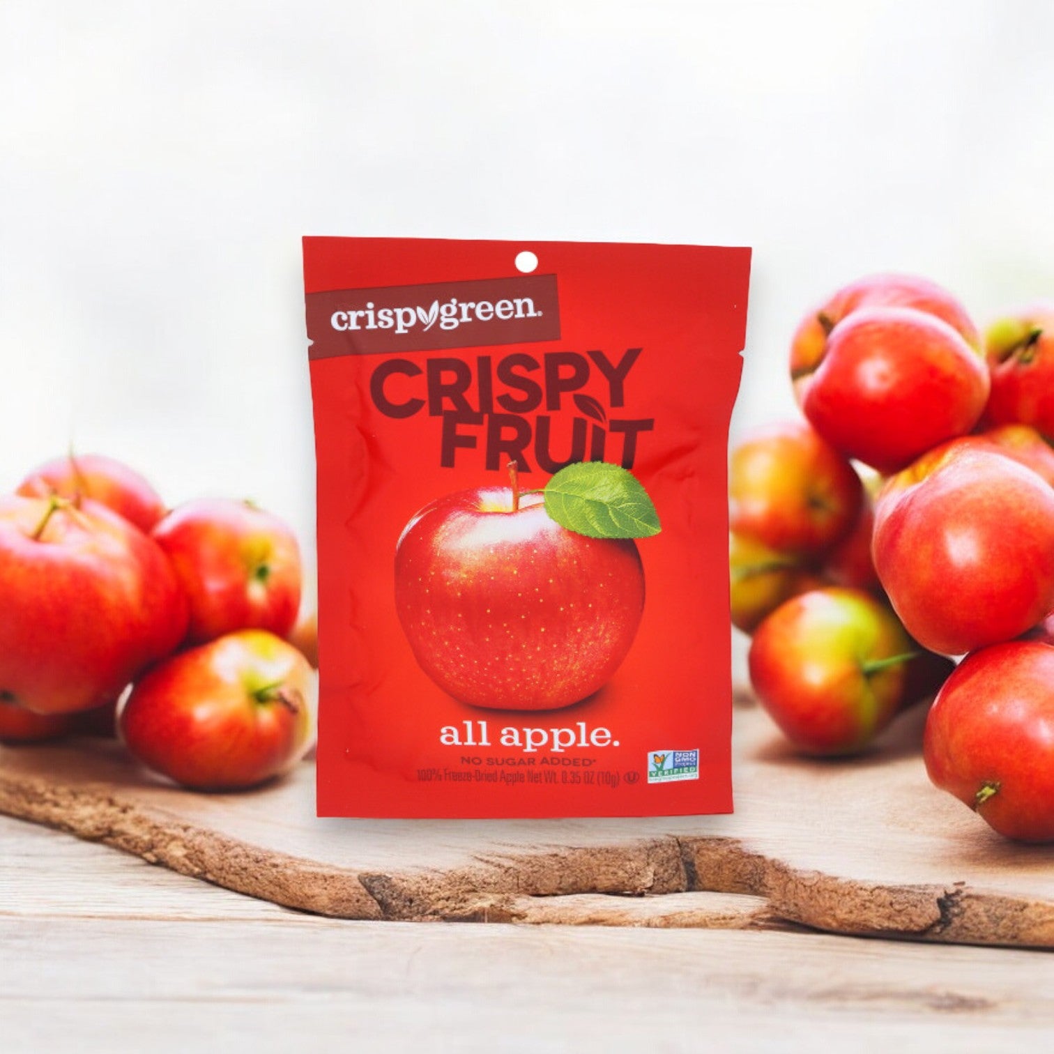 A red bag of Crispy Fruit All Apple-0.35 oz by Crispy Green sits on a wooden surface surrounded by fresh apples, showcasing a delicious and healthy snack made from freeze-dried fruit.