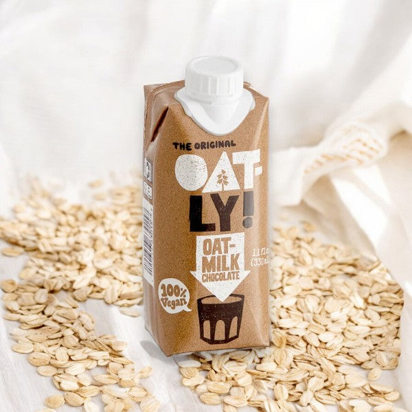 An 11 fl oz carton of Oatly Chocolate Oat Milk is placed on a bed of oats against a white background. The dairy-free carton is labeled "100% Vegan.