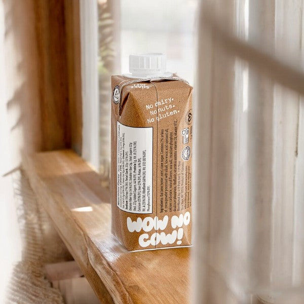 An 11 fl oz carton of Oatly Chocolate Oat Milk sits on a wooden windowsill, sunlight streaming through sheer curtains. This vegan, dairy-free beverage by Oatly highlights that it contains no nuts or gluten.