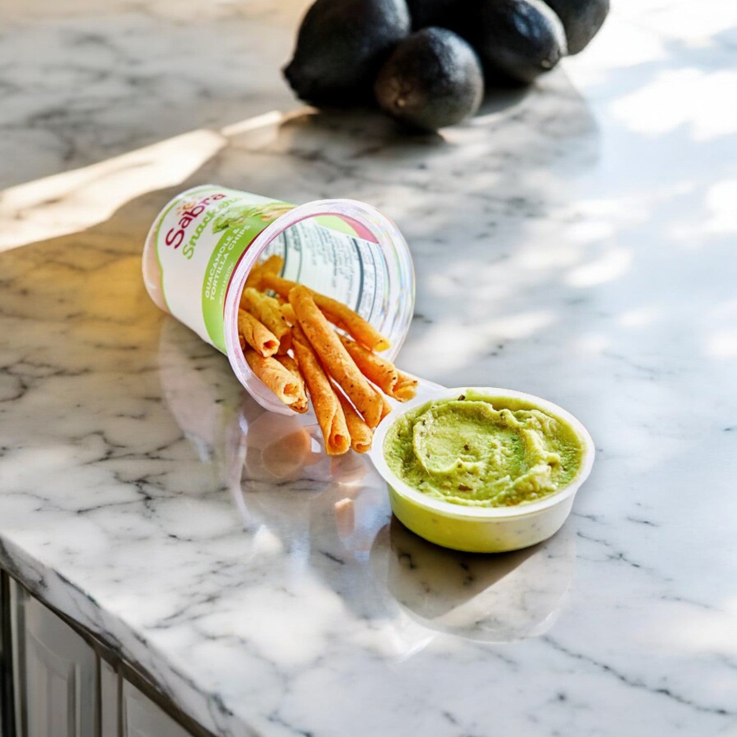 A tub of Sabra Guacamole with Rolled Tortilla Chips, 2.8 oz - 1 Count tipped over beside an open container, resting on a marble countertop with whole Hass avocados in the background.