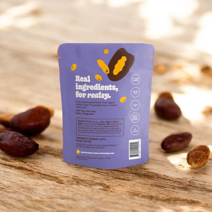 A package of Realsy Peanut Butter Dates with the label "Real ingredients, for realsy." These treats, sourced from a family-owned date farm, showcase several dates and creamy peanut butter scattered around the package on a wooden surface. Perfect for lasting energy throughout your day.