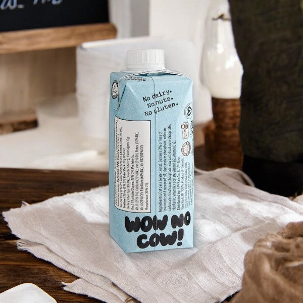 An 11 fl oz carton of Oatly Original Oat Milk, branded with "Wow No Cow!" and described as a dairy-free alternative free from nuts and gluten, sits on a beige cloth on a wooden surface.