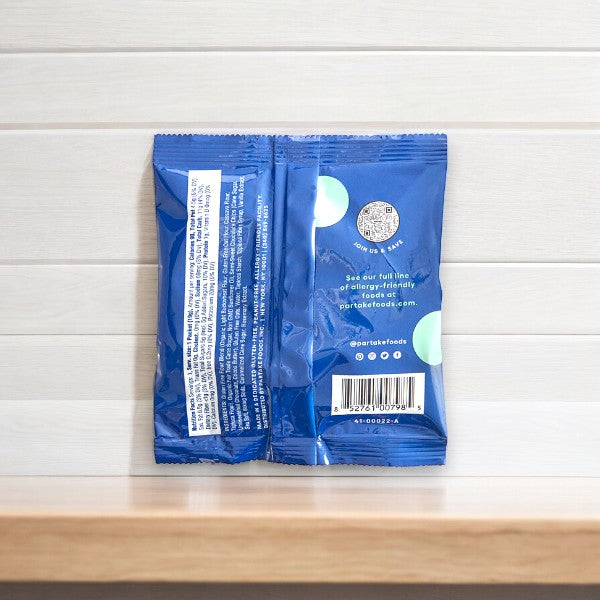 A blue packet labeled "Partake Crunchy Mini Chocolate Chip Cookie Snack Packs, 1 Count" with text and a circular logo on the back, placed against a tiled wall. The text includes website and social media information, highlighting its vegan and allergy-friendly Crunchy Chocolate Chip flavor.