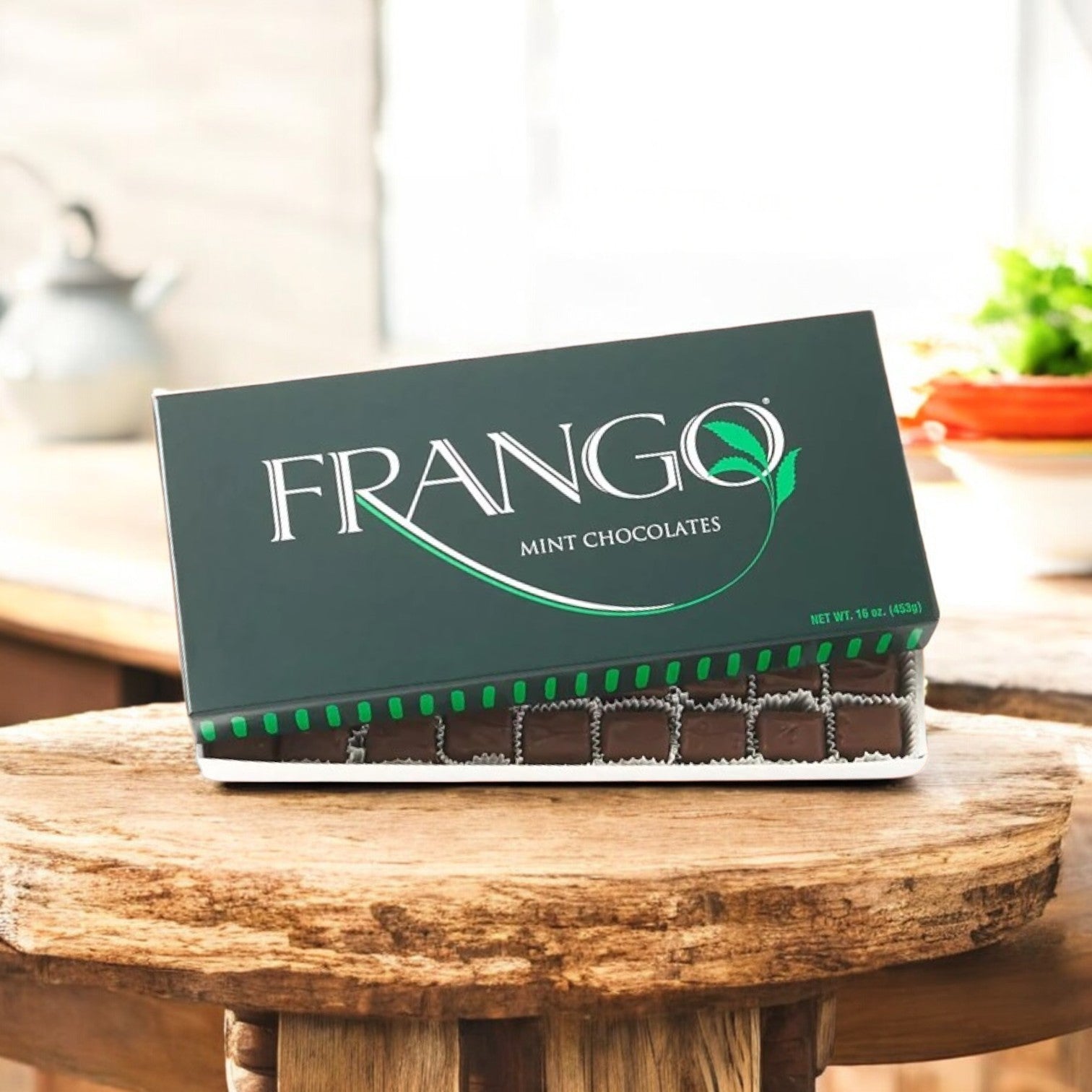 A Holiday Gift Box of Macy's Frango Mint - 1 LB Milk Mint Chocolates sits elegantly on a wooden table, with blurred kitchen items creating a cozy backdrop.