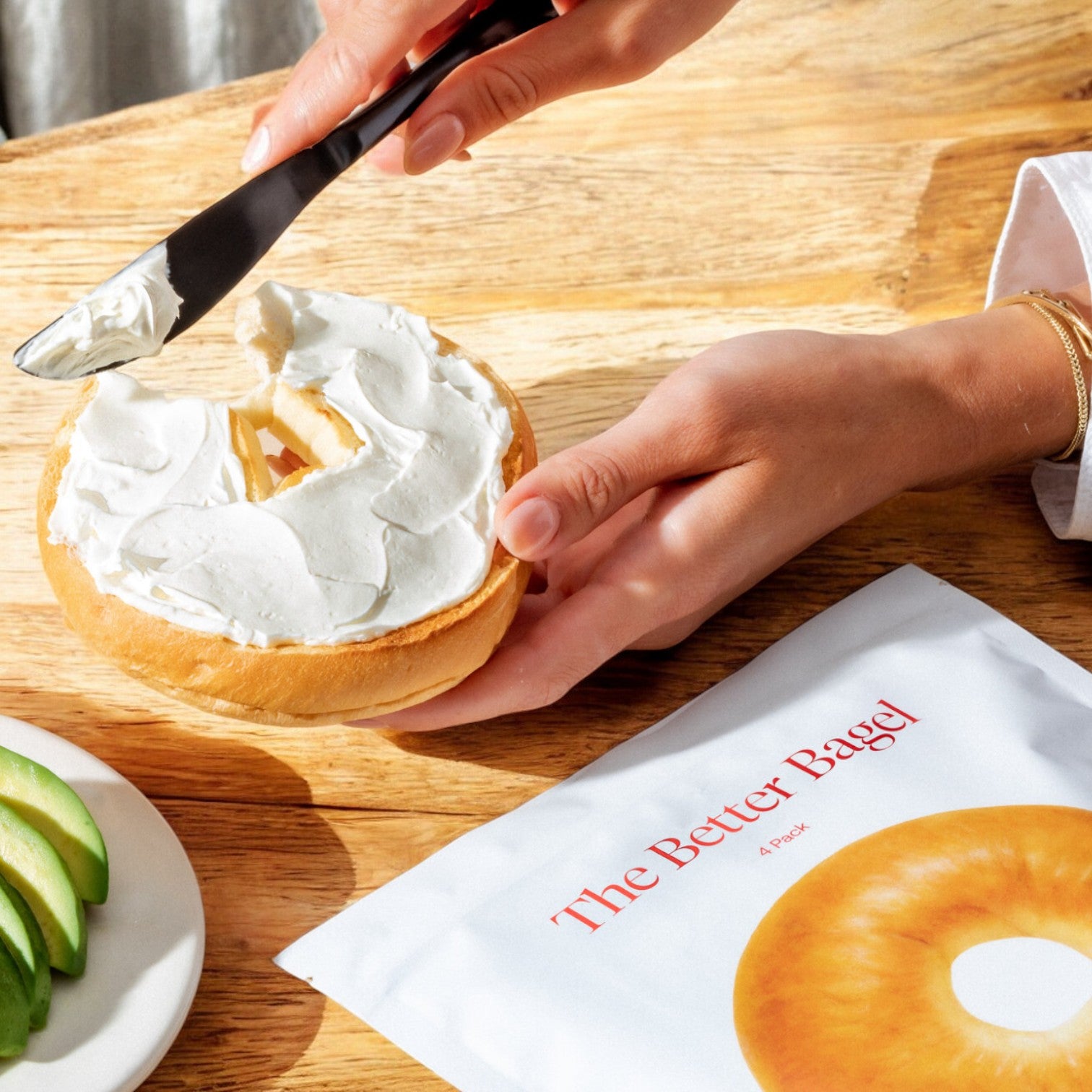 A person spreads cream cheese on a BetterBrand's The Classic bagel using a knife. A packet labeled "Better Brand - The Better Bagel - The Classic- 1 Bag- 4 Bagels Total" and sliced avocados are visible on the wooden table, hinting at the chewy interior that promises satisfaction with every bite.