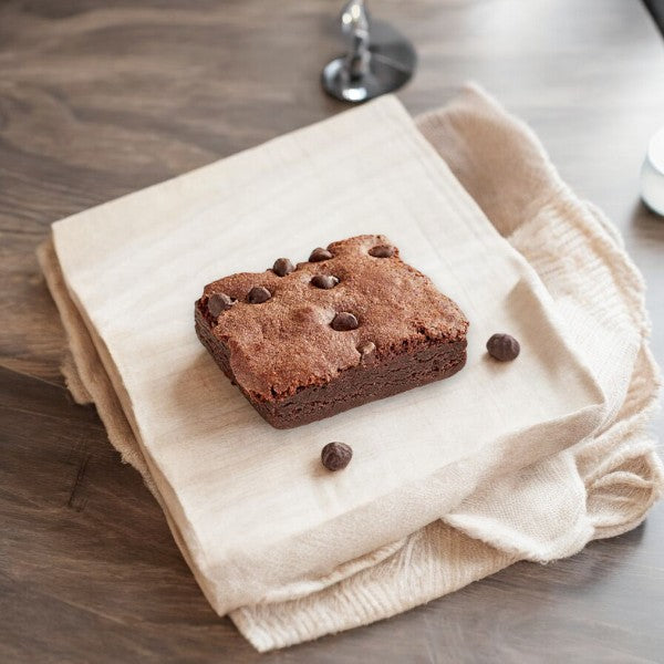 A square, gluten-free **David's Cookies Chocolate Chip Brownie**, weighing 3.5 oz and individually wrapped, placed on a beige napkin over a wooden surface.