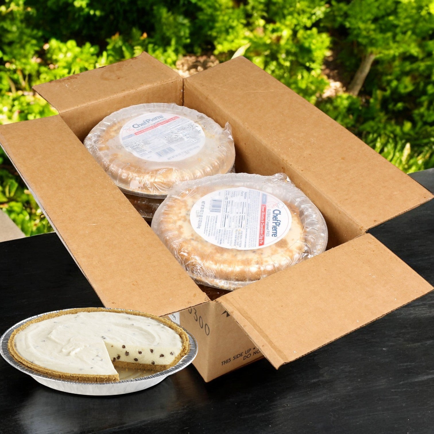 A cardboard box contains three Chef Pierre Sweet Ricotta and Chocolate Chip Pies, featuring a delicious pie next to an open one with a slice cut out on a black table. The backdrop of greenery adds freshness to this Italian-inspired dessert scene.