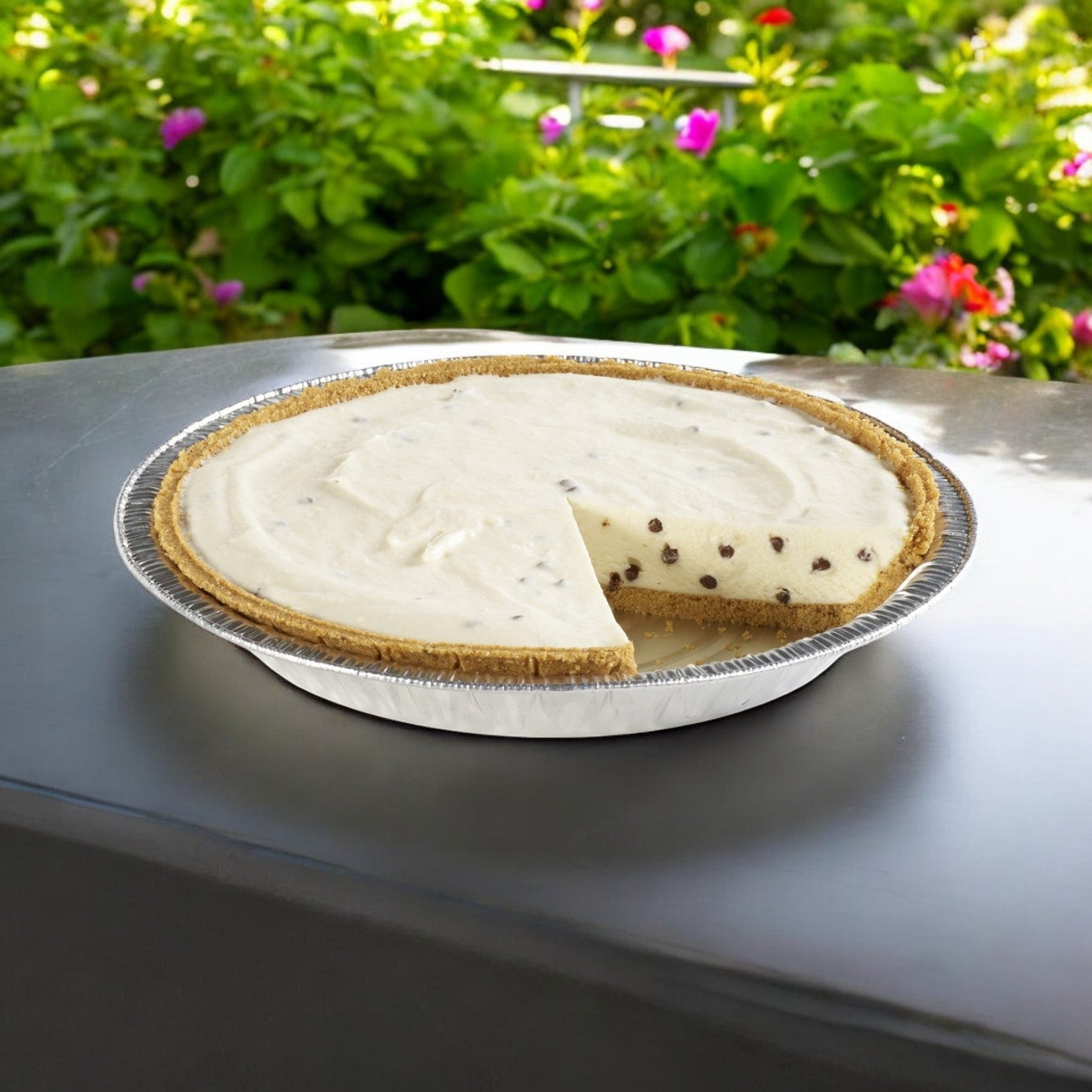 A Chef Pierre Sweet Ricotta and Chocolate Chip Pie rests on a gray surface with a slice missing. In the background, vibrant green foliage and pink flowers enhance the scene with nature's artistry.