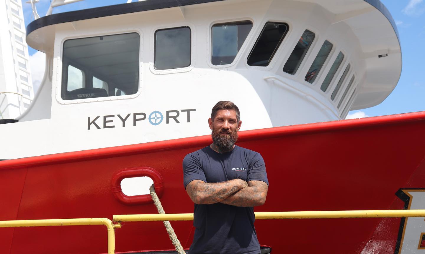 A bearded man stands confidently with arms crossed in front of the Keyport boat, known for its premium Golden Alaskan King Crabs, sustainably sourced and available in a 10-pound box with 16/22 count.