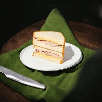 A Taylor Farms Turkey and Cheddar Sandwich, sliced in half, is placed on a white plate with a green napkin underneath, accompanied by a knife.