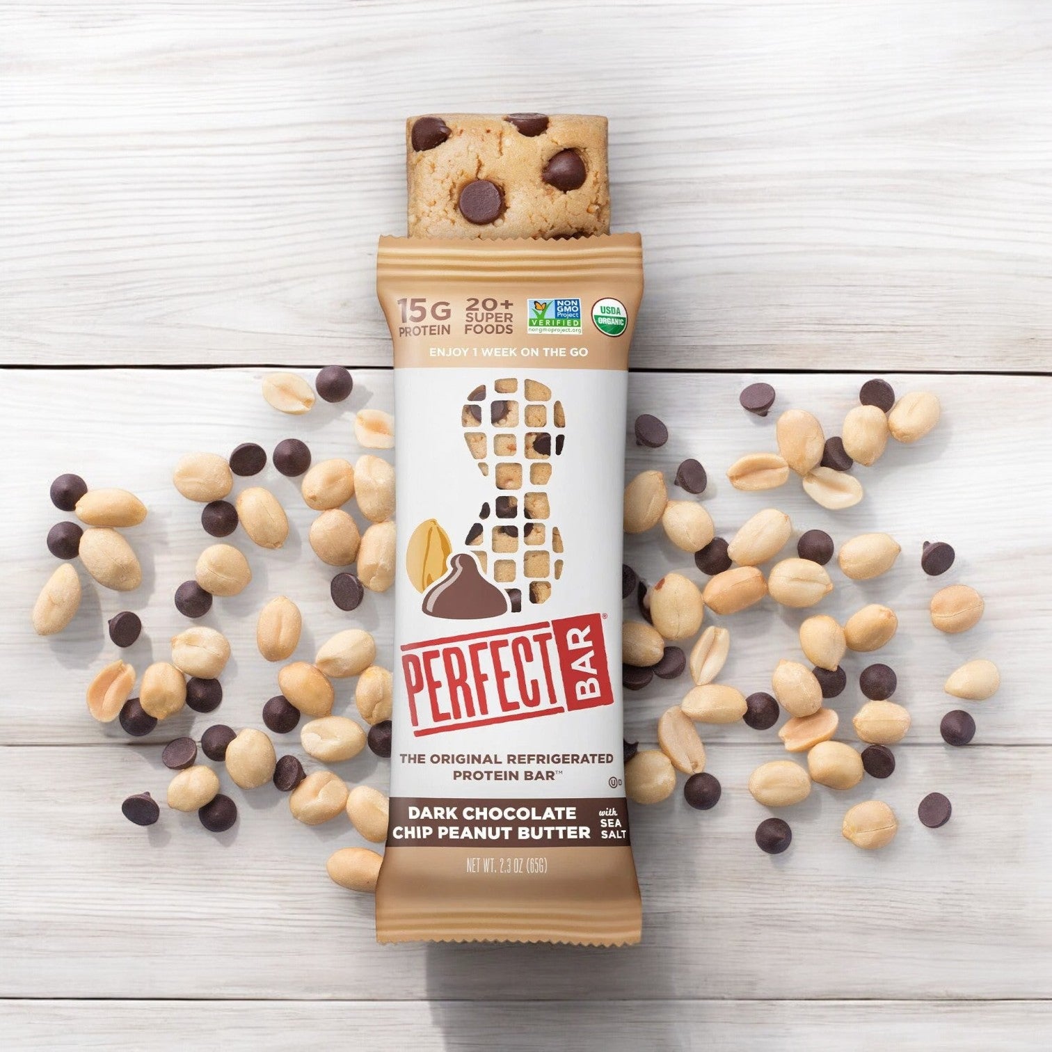 A Perfect Bar Gluten-Free Dark Chocolate Chip Peanut Butter Protein Bar, 2.3 oz - 1 Count, partially unwrapped, surrounded by scattered peanuts and dark chocolate chips on a wooden surface.