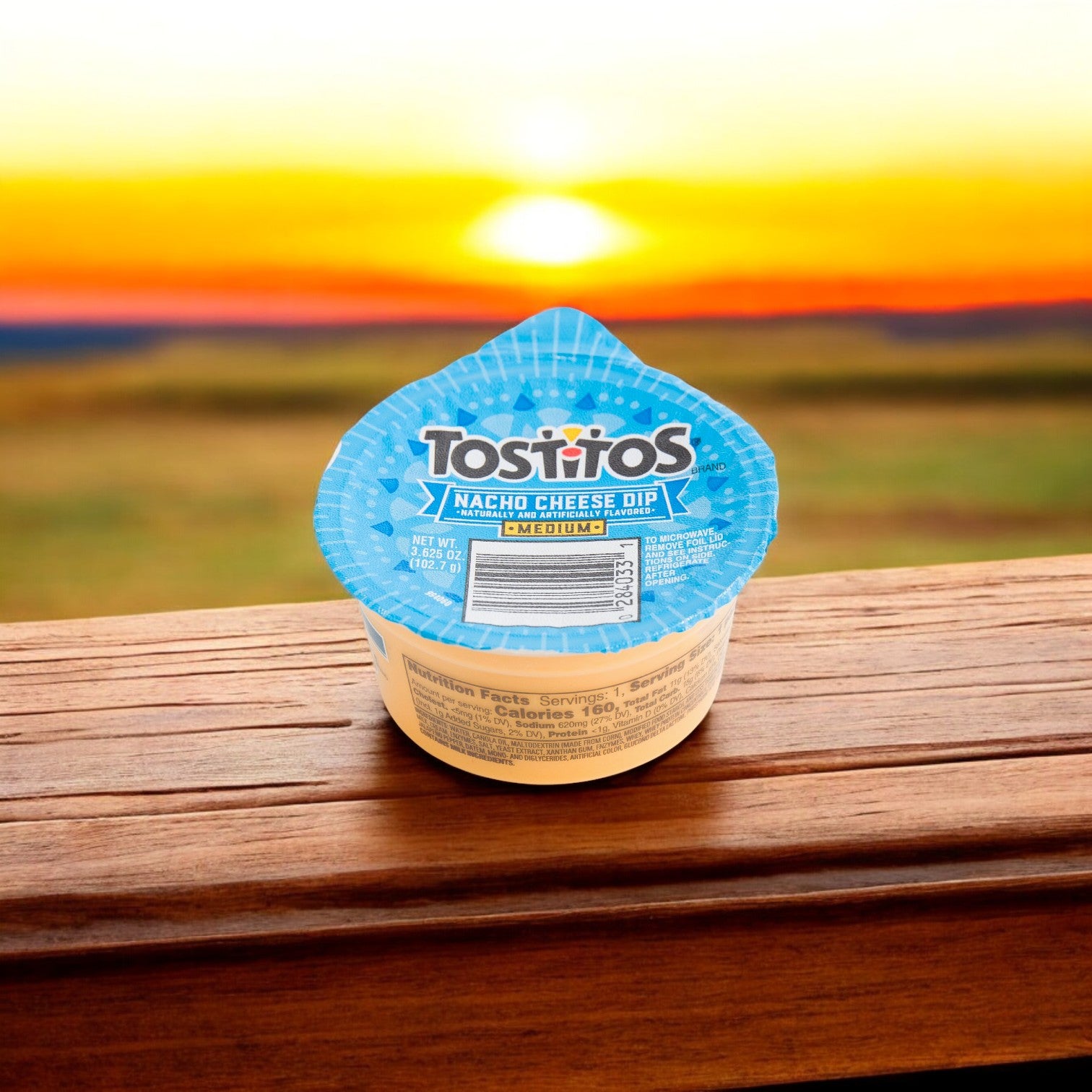 A Tostitos Nacho Cheese Dip Cup from the 30 pack (3.625 oz each) sits on a wooden surface, perfectly paired with Tortilla Rounds, as a vibrant sunset paints the background.