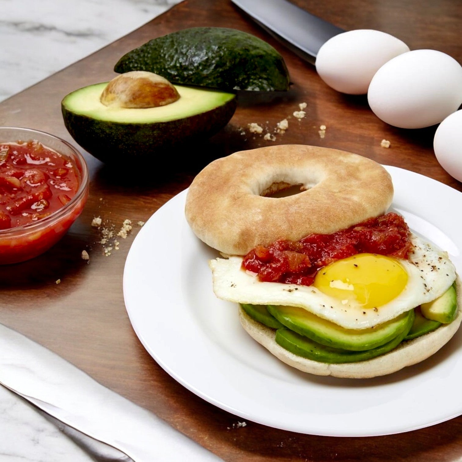 A plate with a bagel sandwich featuring avocado slices, a fried egg, and Tostitos Medium Salsa To-Go Cup alongside an avocado, eggs, a knife, and a bowl of Tostitos Salsa Verde sits on a wooden surface.