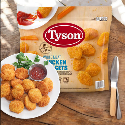 A package of Tyson White Meat Chicken Nuggets, Frozen, 5 lbs, is displayed on a wooden surface. In front, a plate of protein-packed chicken nuggets with a side of ketchup, a fork, and a knife is shown.