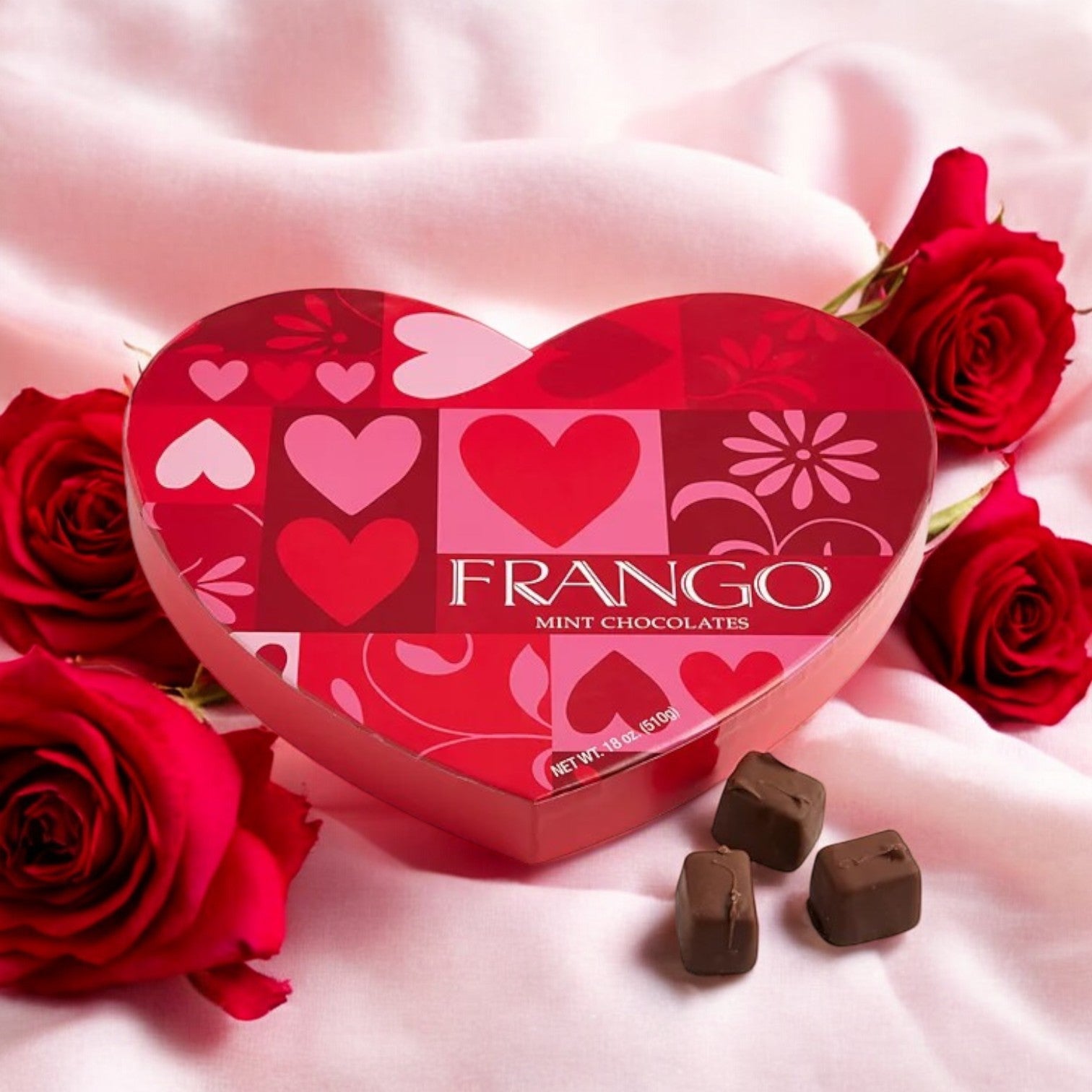 A Macy’s Frango Chocolates Valentine's Day Heart Milk Mint Box of Chocolates (18 oz) is nestled among red roses on a pink fabric background, accompanied by three milk chocolate mint delights.