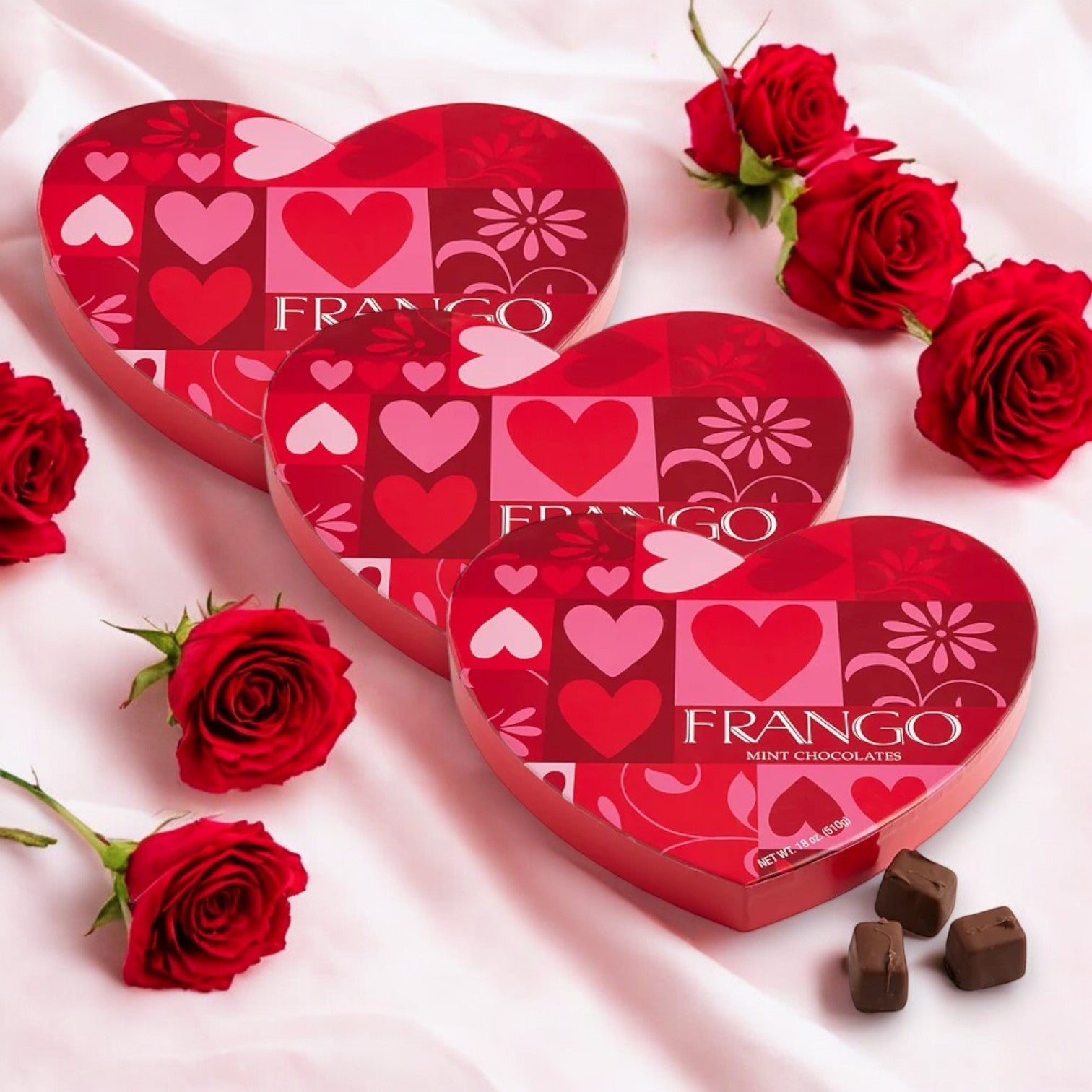 Three boxes of Macy's Frango Valentine's Day Heart Milk Mint Chocolates, 18 oz each, rest on a silky surface adorned with vibrant red roses and scattered chocolate pieces—an ideal indulgence for Valentine's Day.