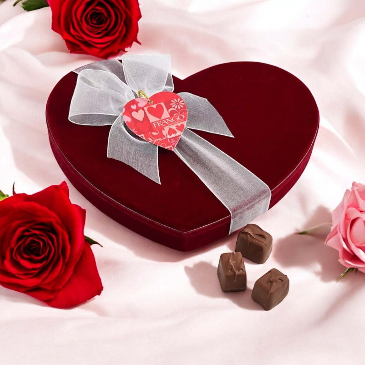 A heart-shaped red box with a white ribbon, surrounded by roses and three chocolates, rests elegantly on pink fabric—Frango Chocolates Valentine's Day Heart Box of Milk Mint Chocolates (14.2 oz), an exclusive charm at Macy's.