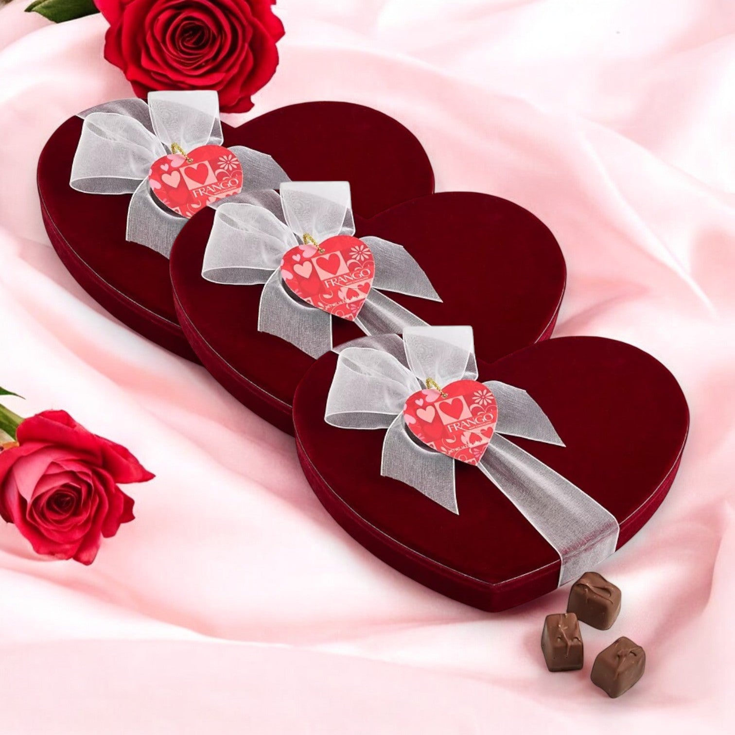 Three Macy's exclusive Frango Chocolates Valentine's Day Heart Boxes with Milk Mint Chocolates, 14.2 oz each, sit on pink fabric surrounded by roses. A delightful setting for a sweet and romantic Valentine’s Day awaits.