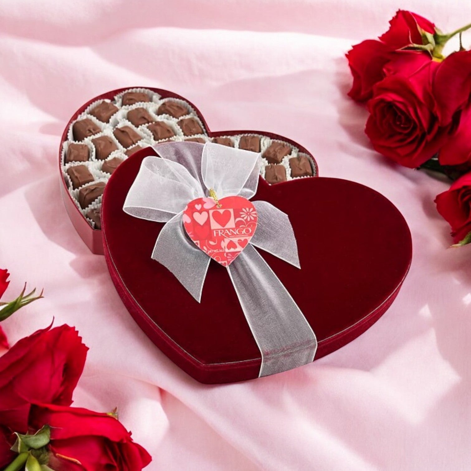 A heart-shaped Macy's Frango Chocolates Valentine's Day box of Milk Mint Chocolates – 14.2 oz, adorned with a white bow and tag, sits among red roses on a pink surface, ideal for Valentine’s Day gifting.