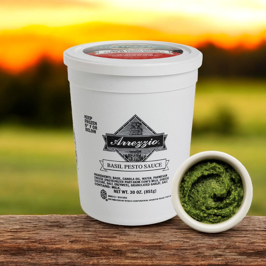 A white container of Sysco's Arrezzio Imperial Basil Pesto Sauce, a delightful Italian blend, rests on a wooden surface with a small bowl of pesto against a blurred sunset backdrop.