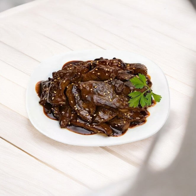 A white plate holding Kevin's Mongolian Beef, featuring tender steak tips glazed in a rich dark sauce and sprinkled with green herbs, is placed on a pale wooden surface.