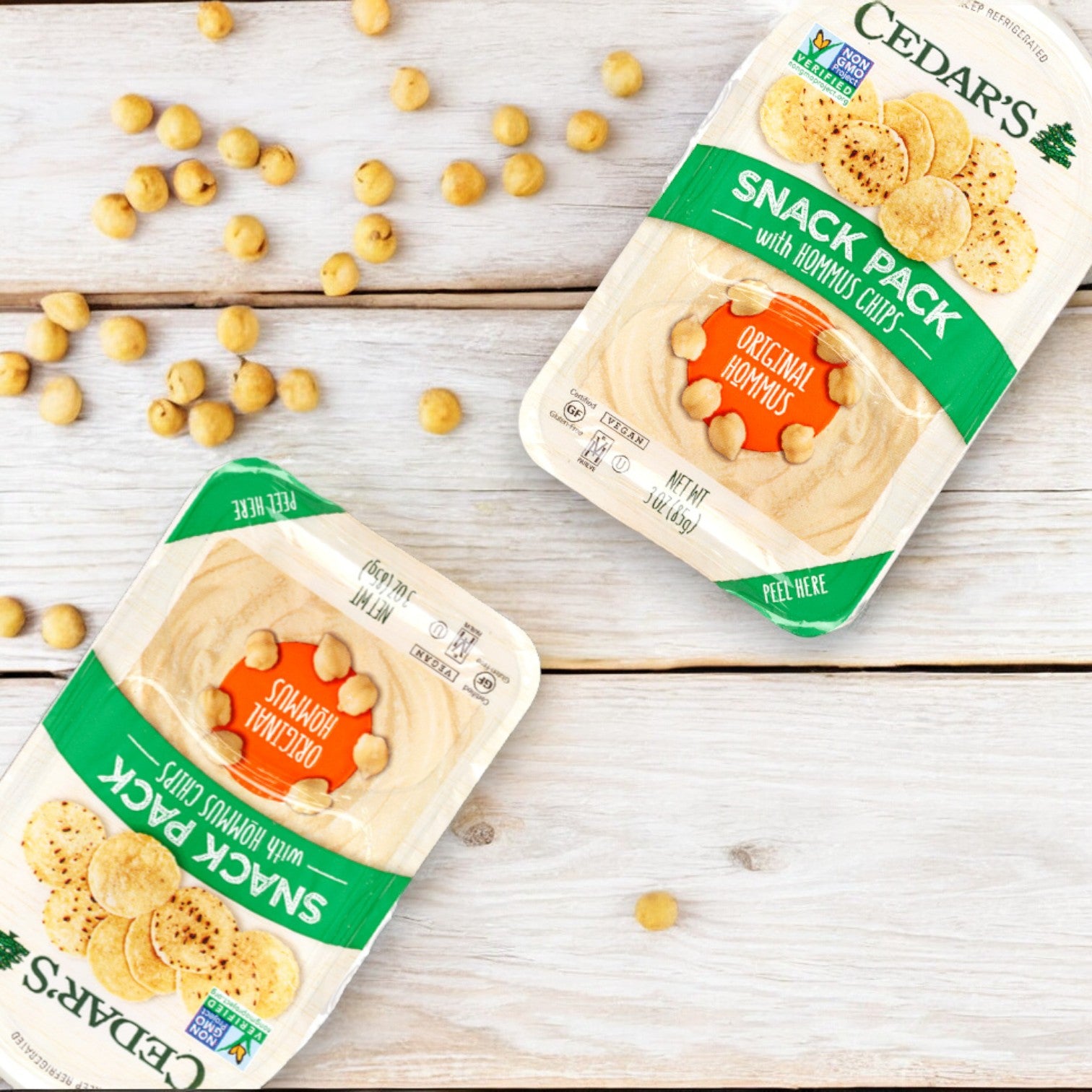 Two packs of Cedar's Snack Pack with Hummus Chips - 1 Count are displayed on a wooden surface, surrounded by scattered chickpeas. The snack packs are labeled "Original" flavor.