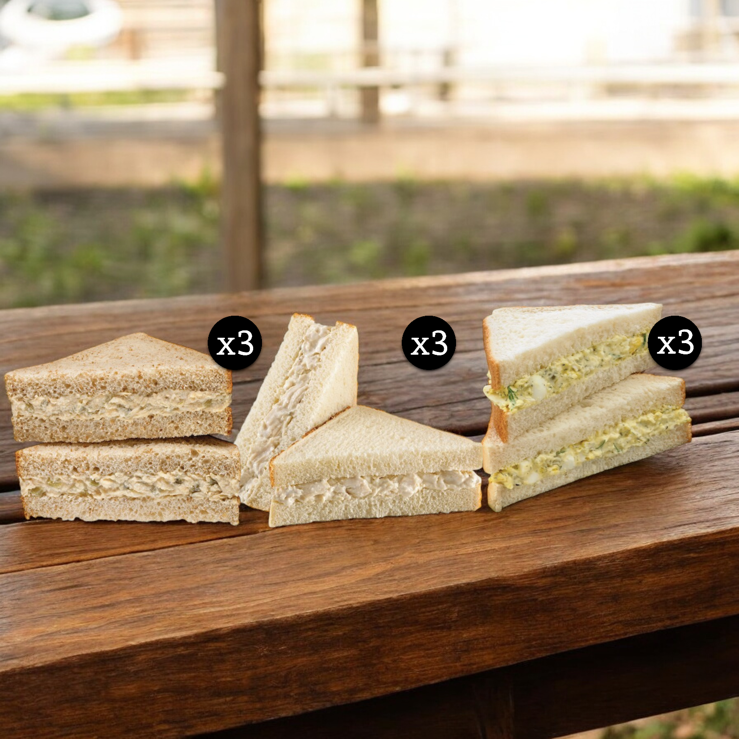 Three sets of sandwiches from the E.A. Sween Deli Salad Variety Pack! are placed on a wooden bench outdoors, with each set containing three triangular chicken salad sandwich pieces.