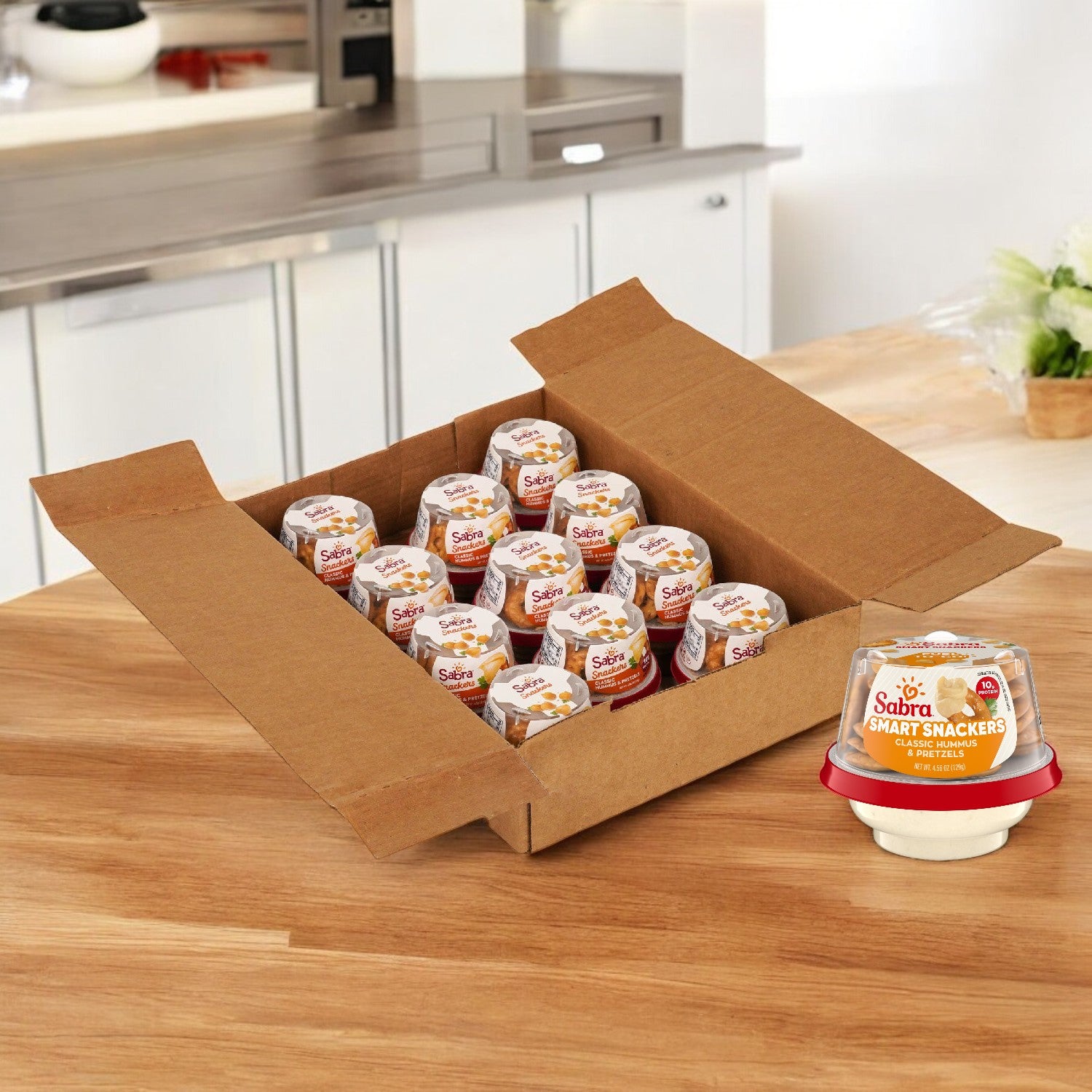 A cardboard box on the kitchen counter holds several Sabra Snackers, including packs of Sabra Classic Hummus with Pretzels, 12 Pack, 4.56 oz each.