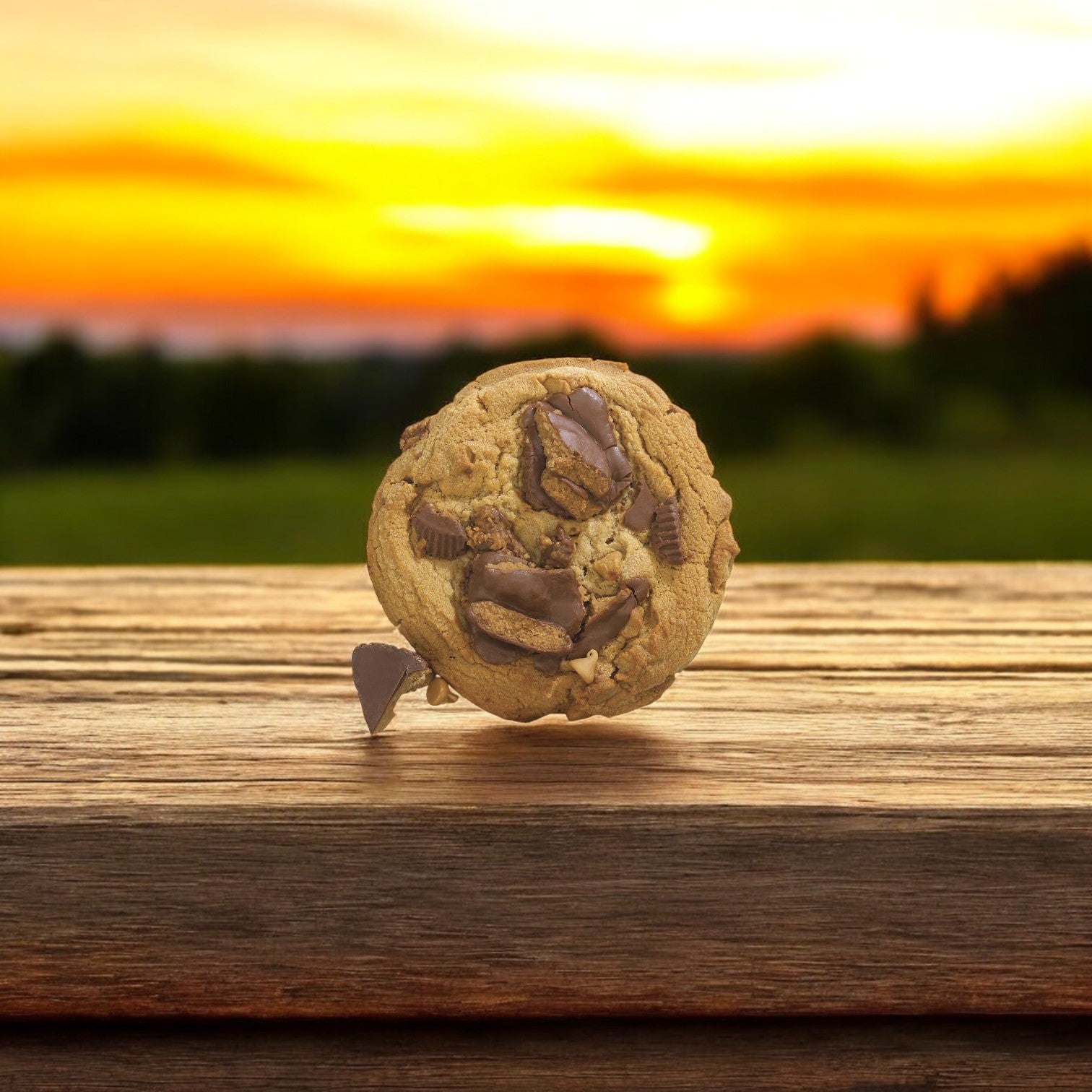 A David's Cookies Dough Cookie - Reese's Peanut Butter Chunk rests on a wooden surface with a blurred sunset in the background.