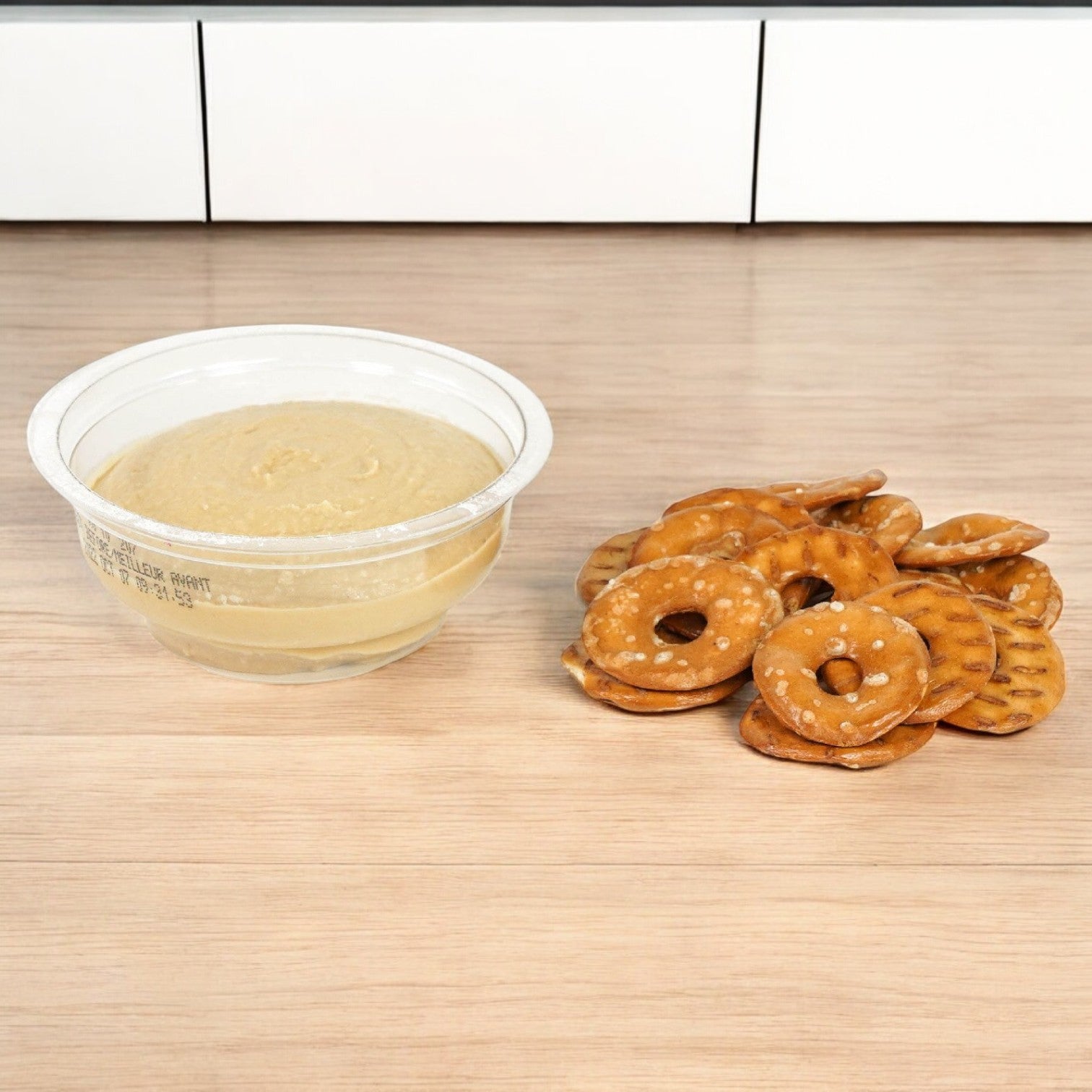 A Sabra Roasted Garlic Hummus with Pretzels snack cup rests on wood, paired with round pretzels.