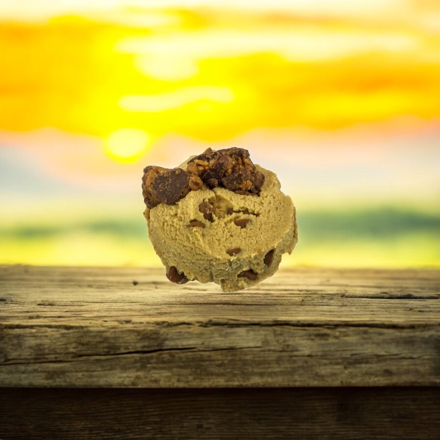 A scoop of David's Cookies Dough Cookie - Reese's Peanut Butter Chunk sits on a wooden surface against the backdrop of a vibrant sunset.