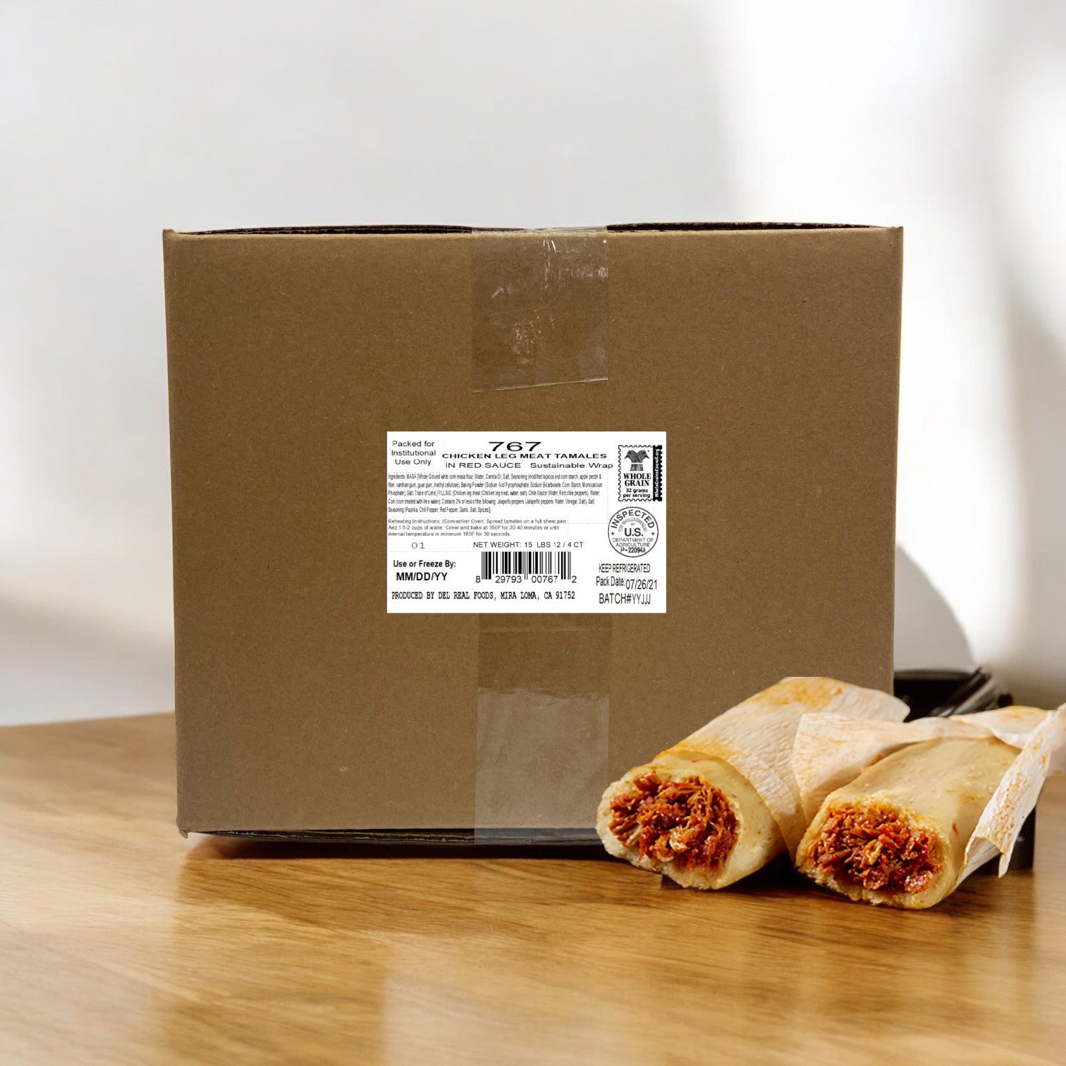 A cardboard box labeled "Del Real Foods Tamale Red Sauce Chicken Whole Grain, 48 Count" sits on a wooden surface next to two unwrapped chicken tamales, highlighting the authentic flavors of Del Real.