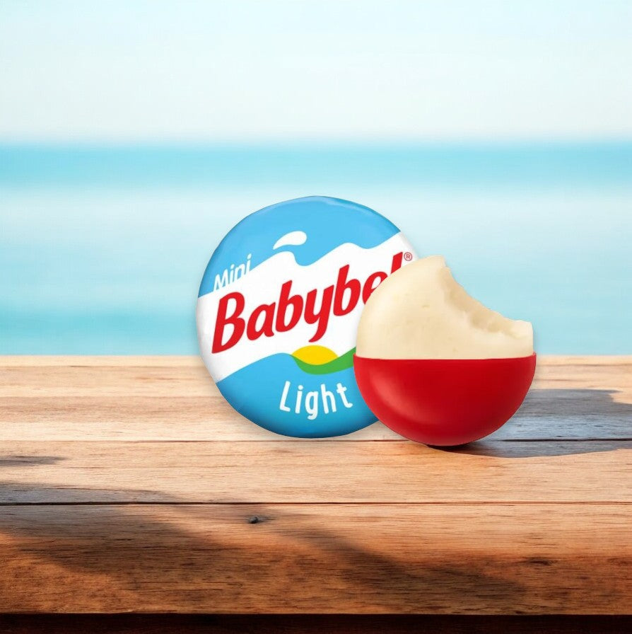 A partially unwrapped piece of Babybel Mini Babybel Light Flavored Snack Cheese, with a bite taken out, rests on a wooden surface against a sea view background, promising an unforgettable snacking experience with real cheese.