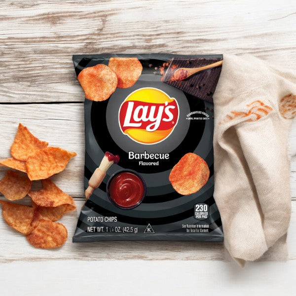 A bag of Lay's® Barbecue Flavored Potato Chips Snack Chips 1.5 oz from Frito Lay is displayed on a wooden surface, accompanied by some chips and a small dip bowl of barbecue sauce, alongside a white fabric napkin with an orange design. This shelf-stable snack offers the perfect combination for your next gathering or solo indulgence.