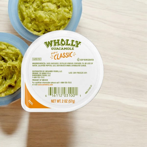 A container of WHOLLY Guacamole Classic Minis, 2 oz - 1 Count, by Wholly, is shown. The lid lists ingredients and details, with two more containers partially seen in the background. This convenient snacking option features hand-scooped Hass Avocado for fresh flavor.