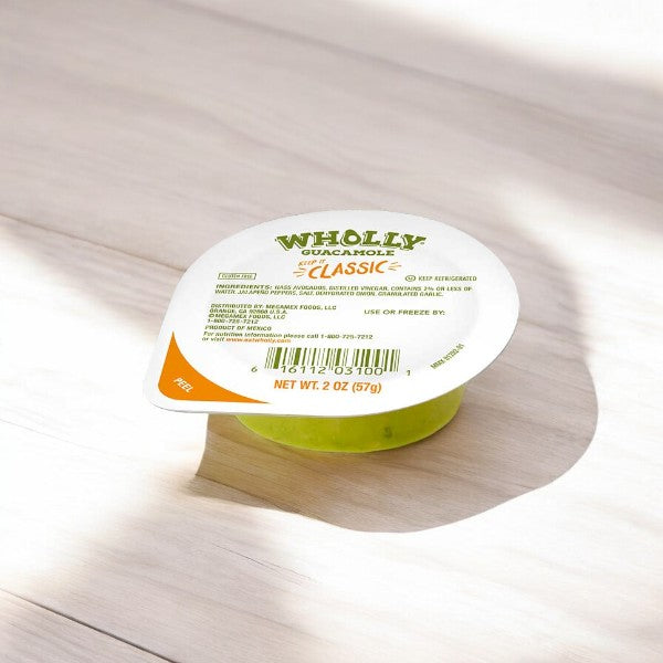 A small plastic container of WHOLLY Guacamole Classic Minis on a light wooden surface. Perfect for convenient snacking, the label provides nutritional information, and the weight of the hand-scooped Hass Avocado guacamole is 2 oz (57g).