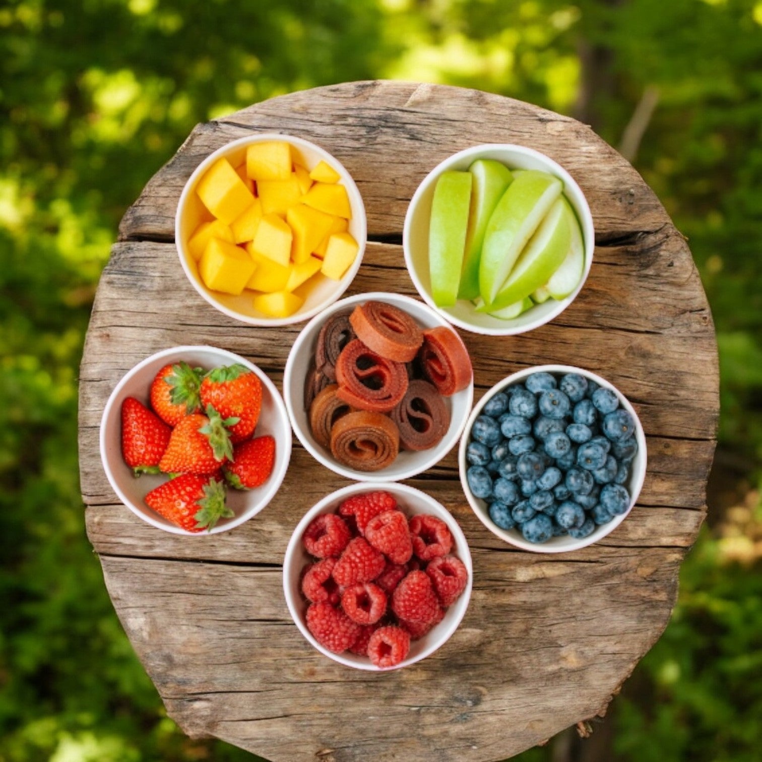 Five bowls of assorted fruits and 100% natural fruit snacks are arranged on a rustic wooden surface. The bowls contain cubed mango, sliced green apple, strawberries, BEAR Raspberry Fruit Rolls (0.7 oz) with no added sugar, blueberries, and raspberries.