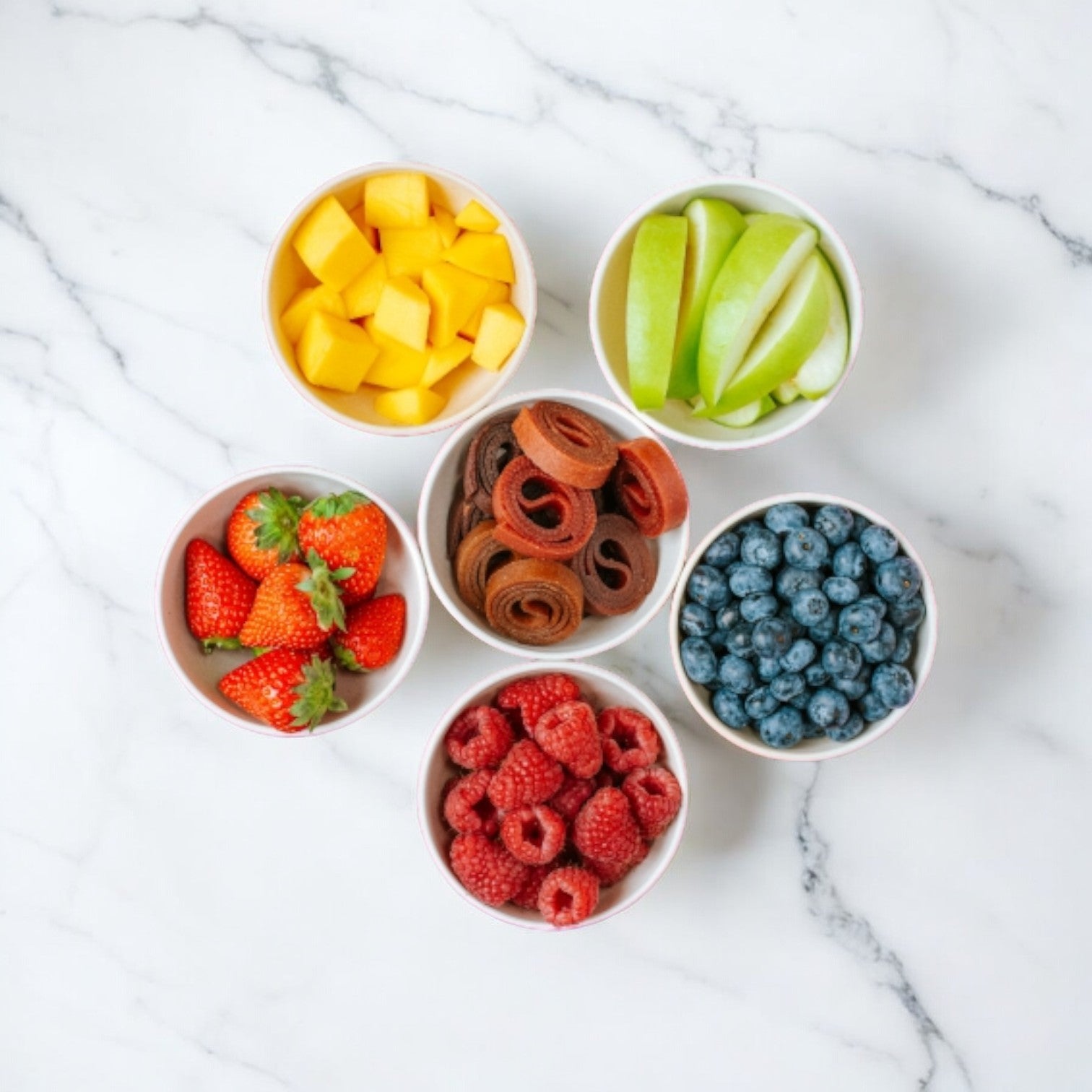 Five small bowls containing diced mango, green apple slices, blueberries, raspberries, strawberries, and Bear Fruit Rolls - Strawberry (0.7 oz) arranged on a white marble surface. These vibrant and healthy snacks from Bear are perfect for a natural fruit snack spread.