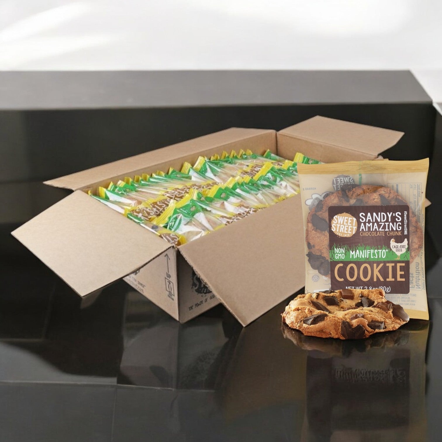 A box of Sweet Street, Sandy's Amazing Chocolate Chunk Manifesto Cookies (48 Count) rests on a glossy black surface, with one individually wrapped 2.8 oz. package and cookie displayed for freshness.