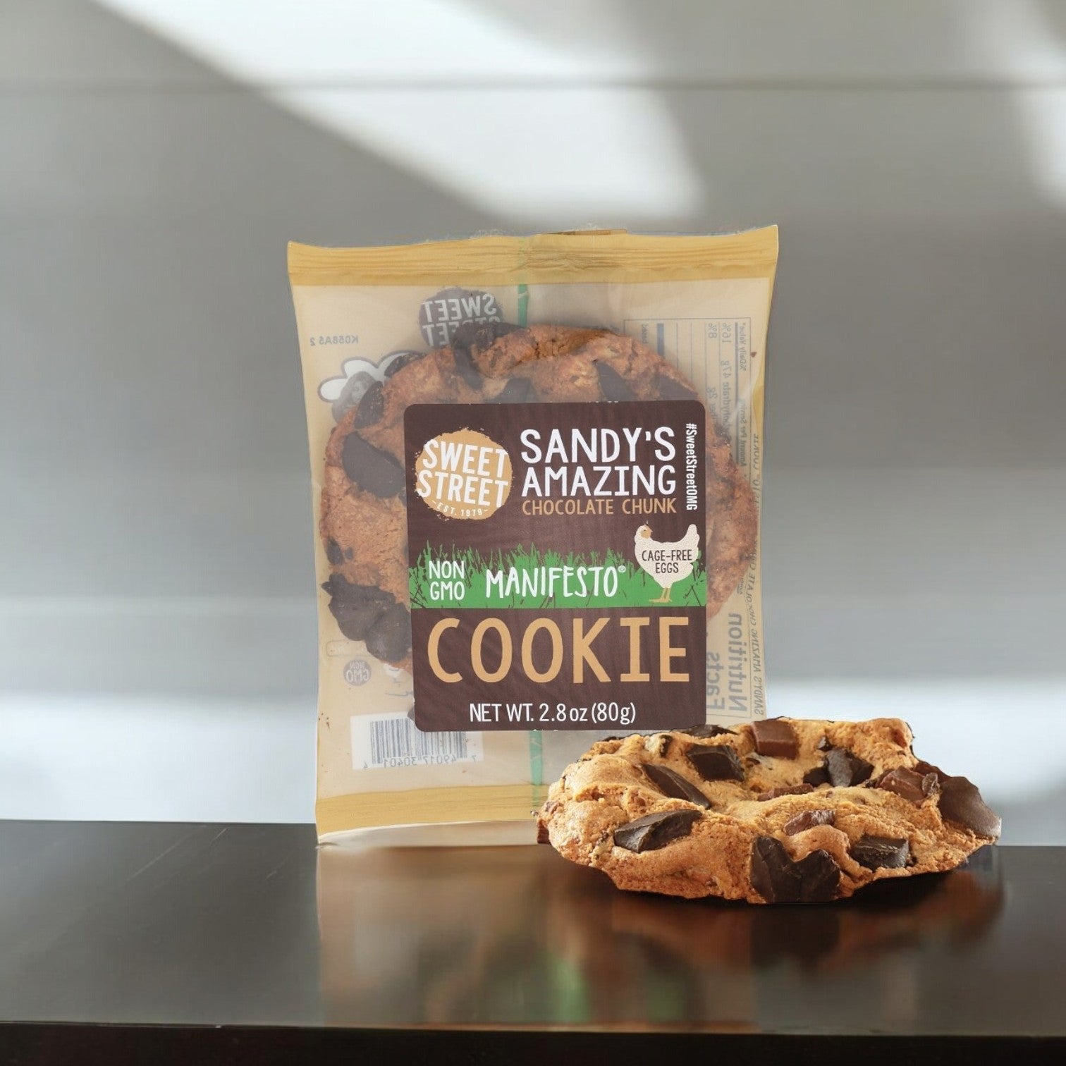 A packaged Sweet Street, Sandy's Amazing Chocolate Chunk 2.8 oz. Manifesto Cookie with another cookie in front on a dark surface showcases Non-GMO Cookies excellence.