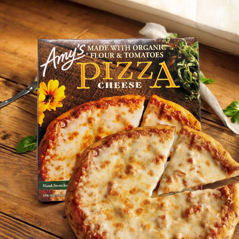 A box of Amy's Cheese Frozen Pizza-1 Count is shown in the background with a cooked vegetarian pizza, featuring one slice slightly removed from the whole, in the foreground. The packaging states it is made with organic ingredients.