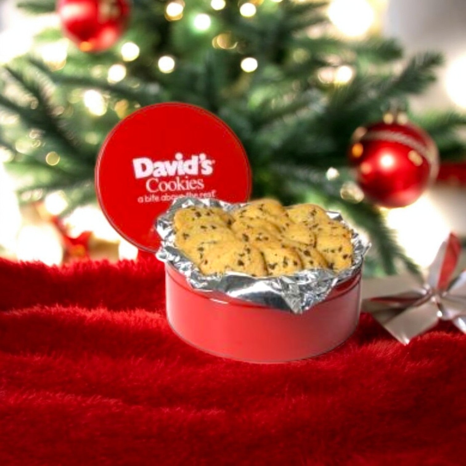 A red tin filled with freshly baked David's Cookies - Chocolate Chip Cookie Dough cookies, each weighing 1.5 oz., sits open on a vibrant red surface, exuding warmth and sweetness in front of a beautifully decorated Christmas tree adorned with twinkling ornaments.