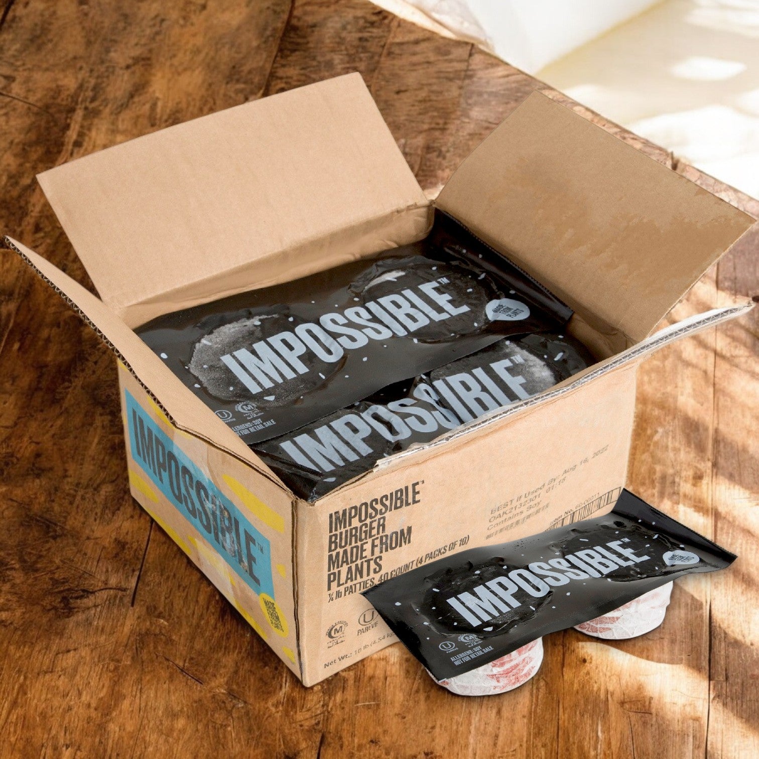 A cardboard box on the wooden floor reveals packaged Impossible Foods plant-based burger patties, made with non-GMO ingredients: Impossible Burger 1/4 Pound Frozen (40/4 OZ).