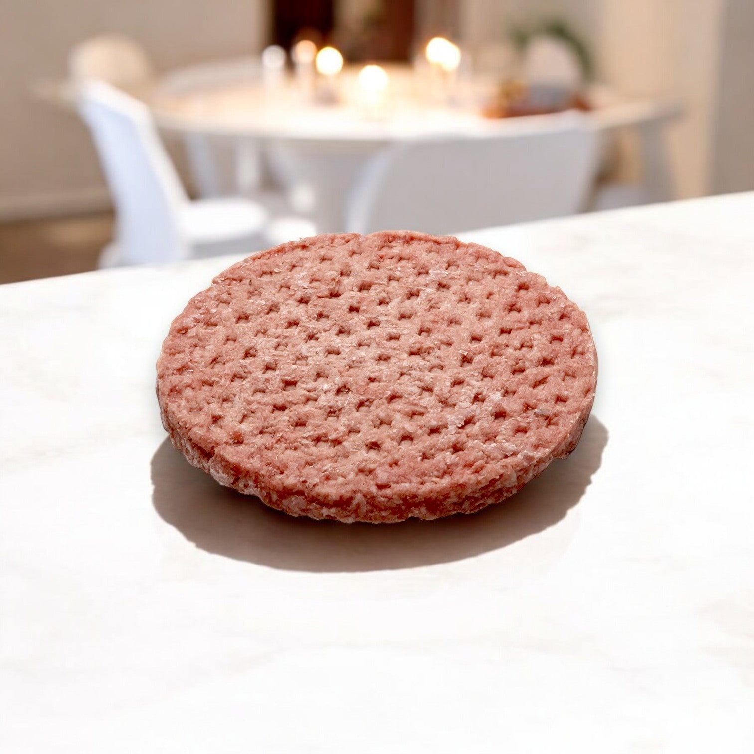 A Fire River Farms Imperial halal beef patty with a 75/25 lean-to-fat ratio sits uncooked on white marble, against a blurred backdrop of table and chairs.