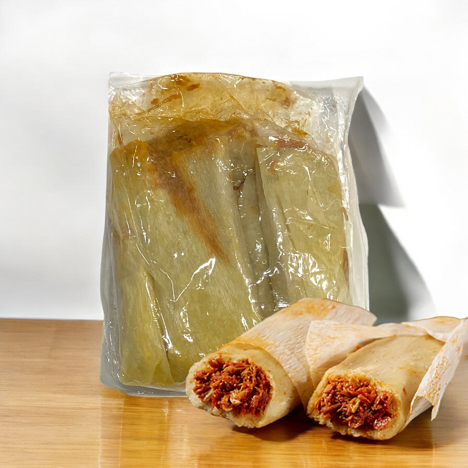 A vacuum-sealed package of Del Real Foods Tamale Red Sauce Chicken Whole Grain, 48 Count, showcasing authentic Mexican flavors, is displayed on a wooden surface.