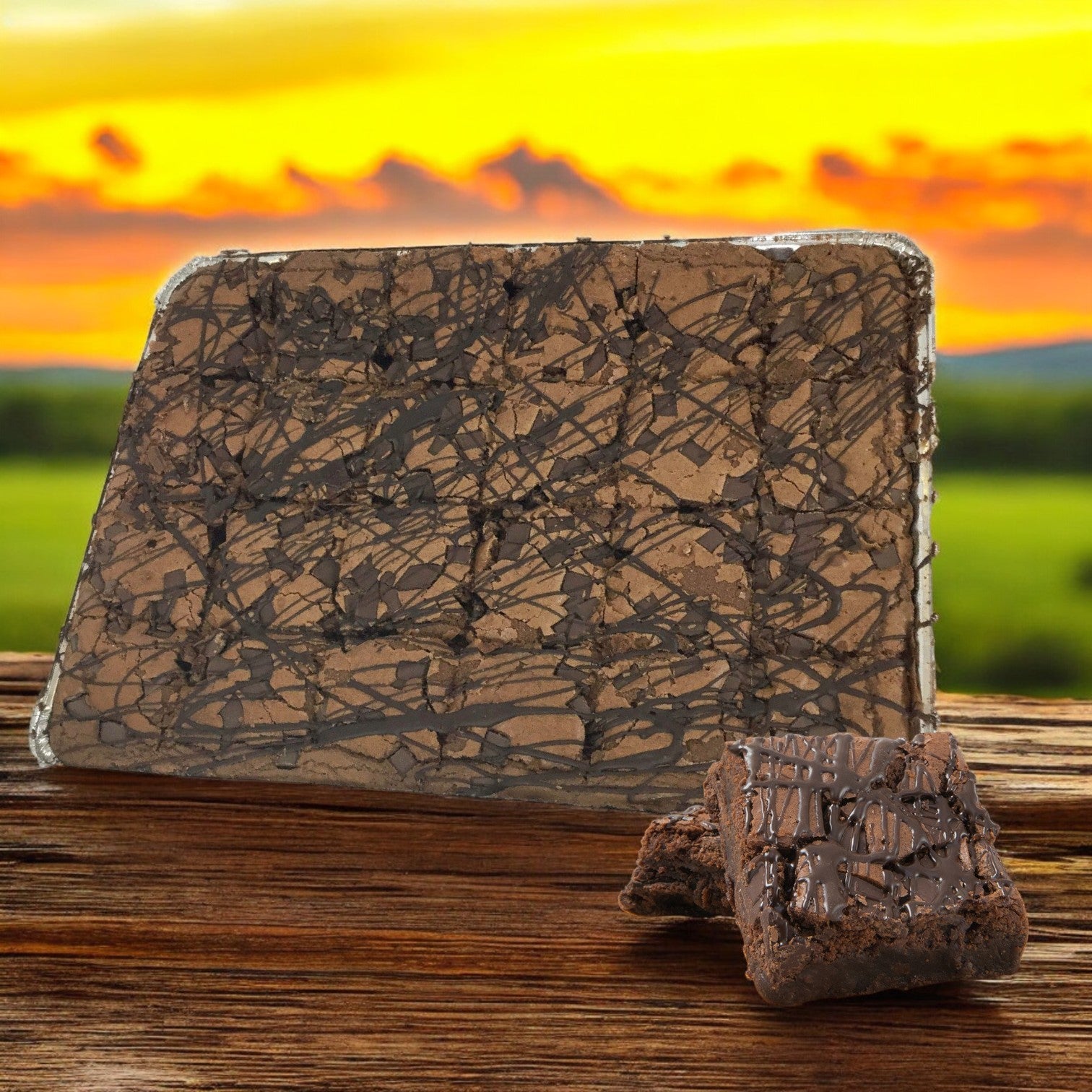 A large slab of David's Cookies Double Callebaut Fudge Brownies with a drizzled pattern, accompanied by two bakery-style cookies and smaller chocolate pieces, set on a wooden surface against a sunset landscape.