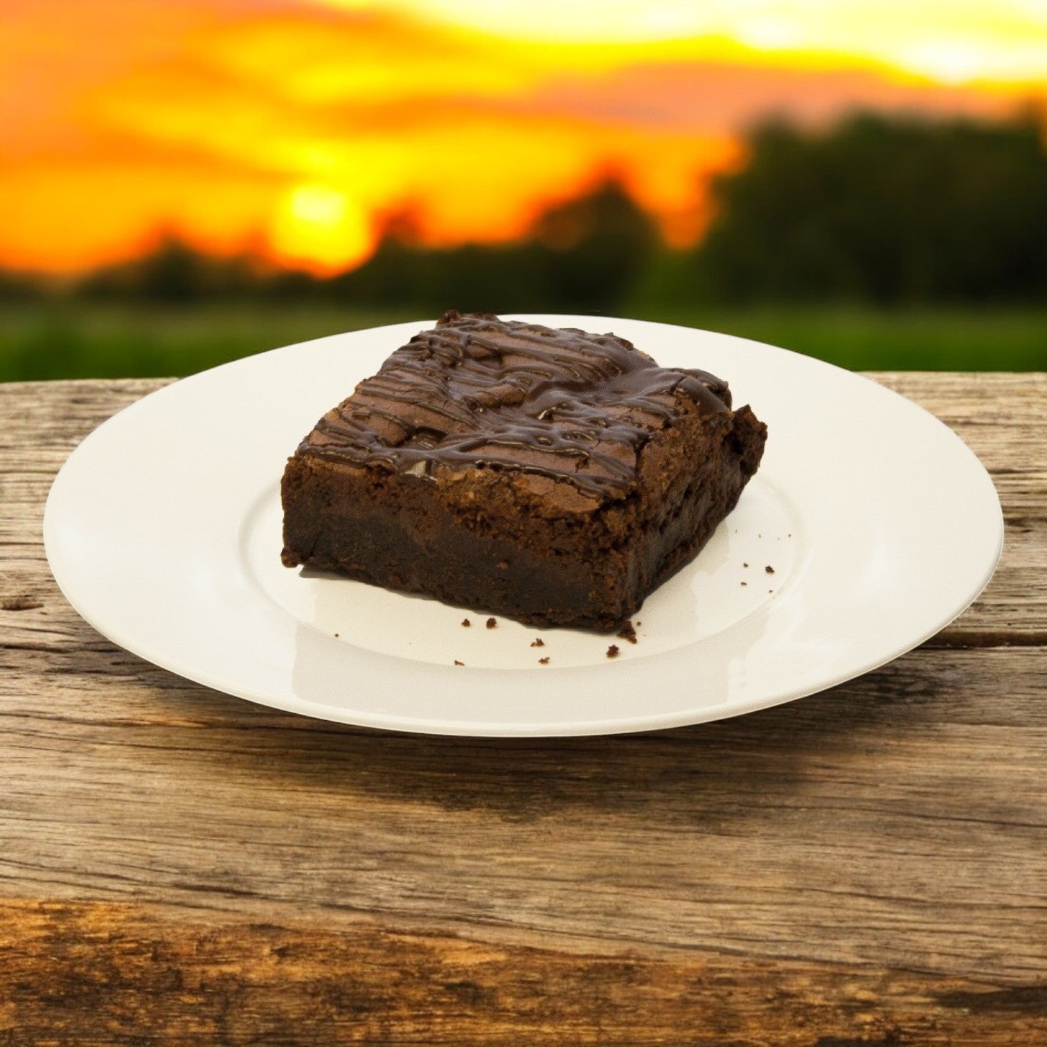 A David's Cookies - Double Callebaut Fudge Brownie rests on a white plate atop a wooden table, framed by a sunset and greenery, capturing the serene pleasure of enjoying fresh bakery-style treats from David's Cookies.