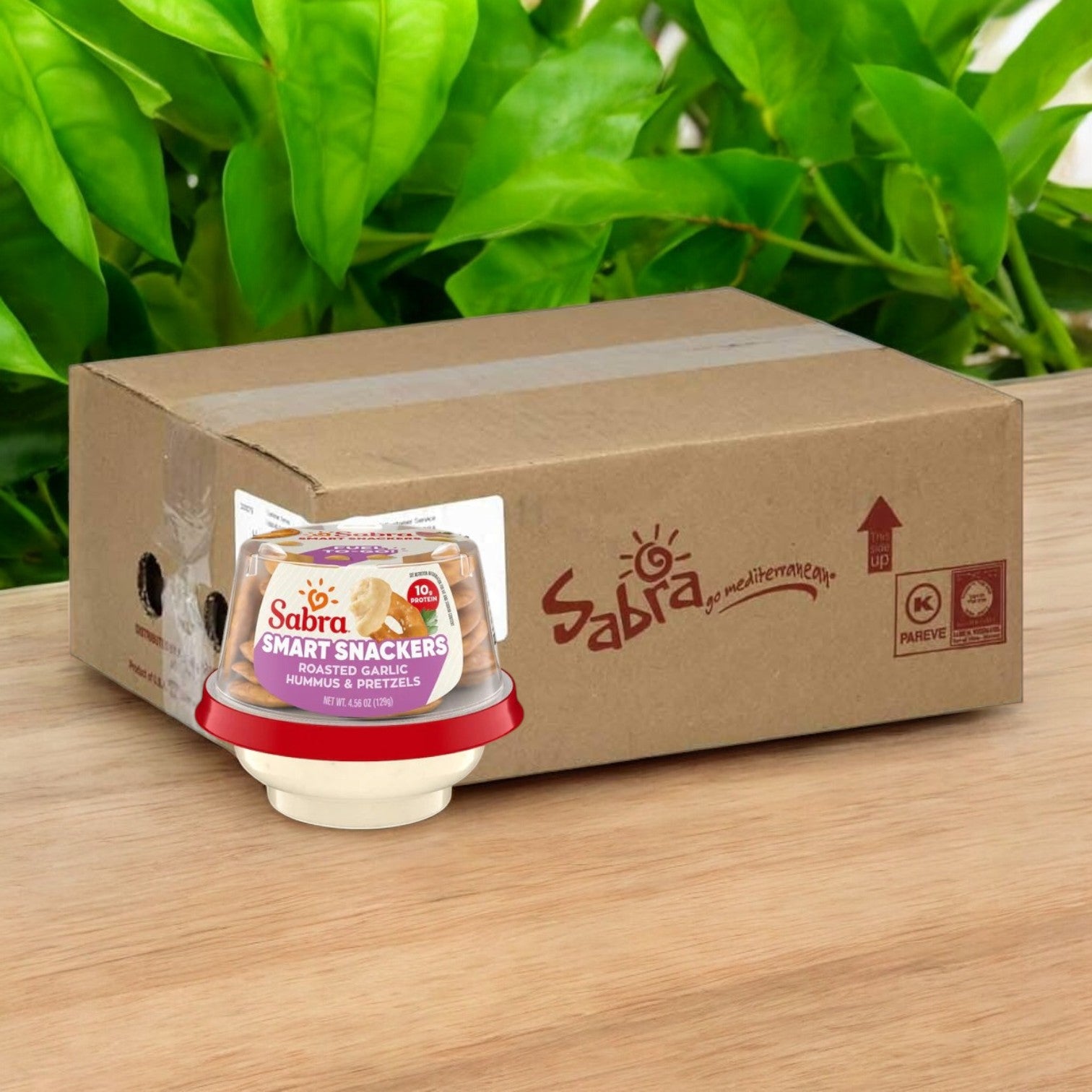 A small container of Sabra Roasted Garlic Hummus with Pretzels sits on a wooden surface, surrounded by green leaves, in front of a Sabra-branded box. This portable snack cup from the 12 pack case is perfect for an on-the-go treat.