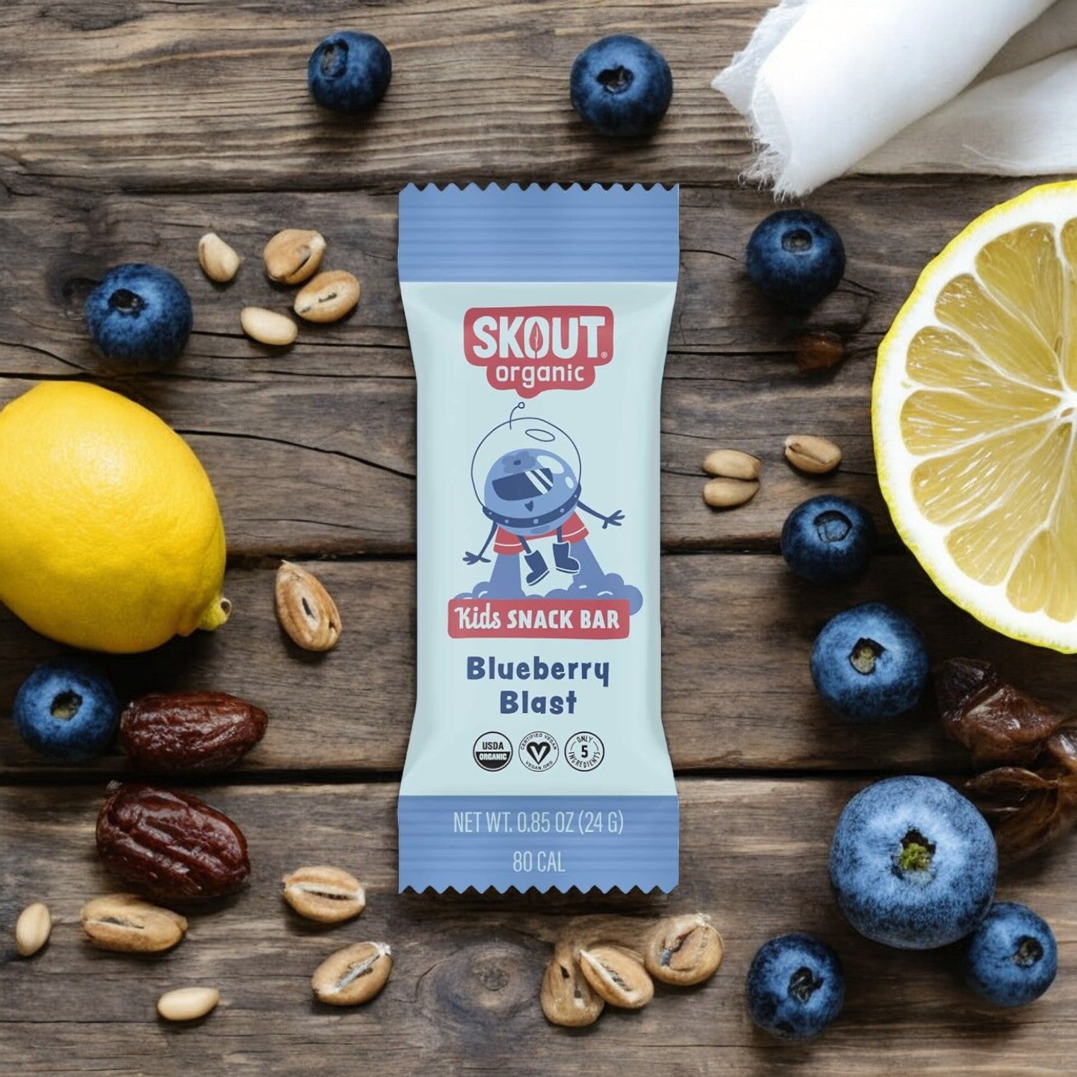 A Skout Organic Snack Bar, Blueberry Blast flavor for kids, is placed on a wooden table surrounded by blueberries, dates, nuts, and halved lemons. Made with plant-based ingredients, it offers a delicious and wholesome treat for kids.