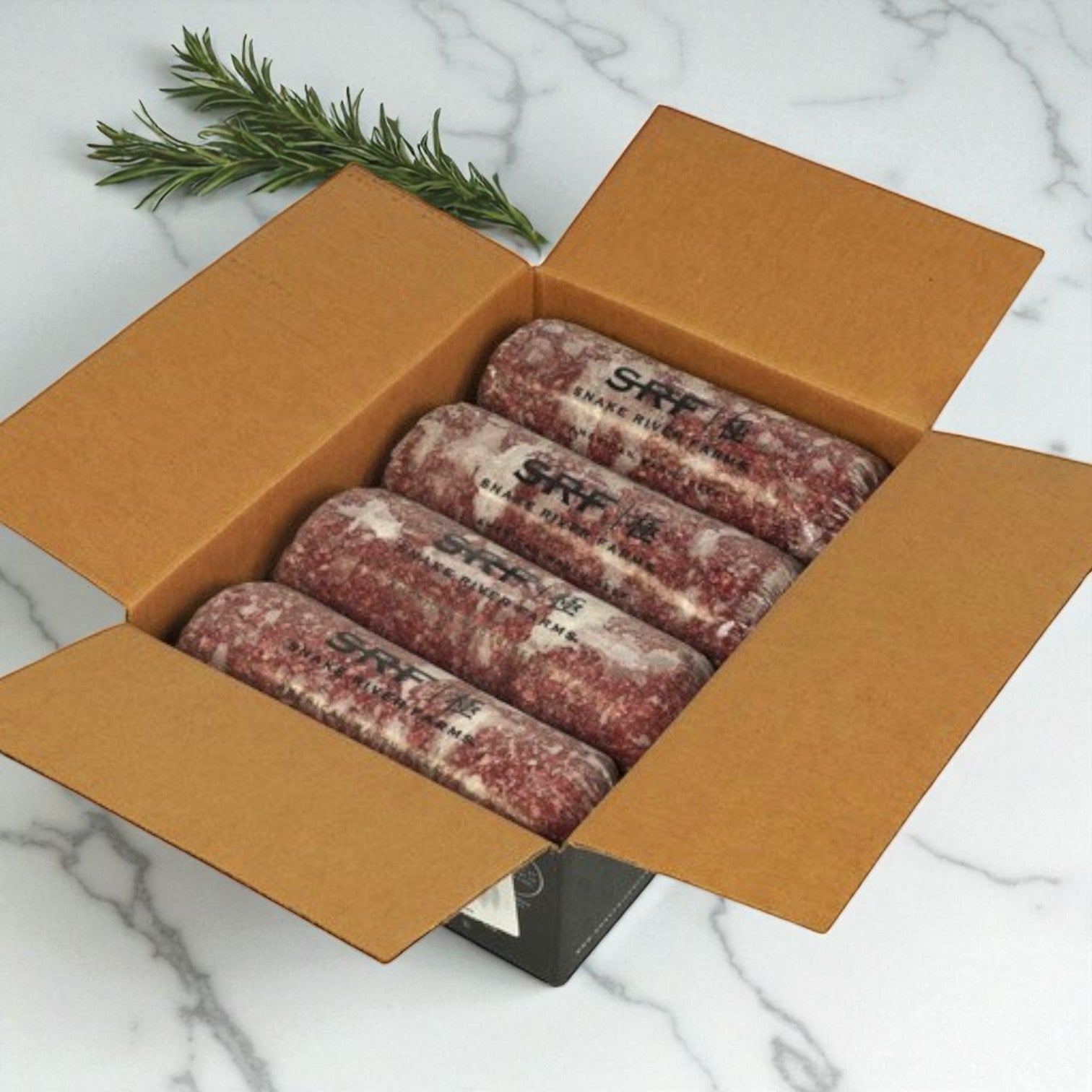 Open cardboard box with four vacuum-sealed rolls of Restaurant Depot’s Snake River Farms Beef Ground – American Style Wagyu 75% Bulk (4/5 lb avg) on a marble countertop, garnished with a sprig of rosemary.